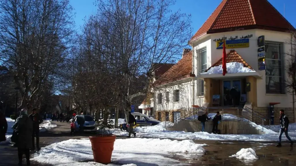 A picturesque winter scene in Ifrane, Morocco, showcasing the charming architecture of the town with its European-inspired buildings. Snow blankets the rooftops and streets, while evergreen trees surround the area. The sky is a clear blue, contrasting with the white snow, and a few people are enjoying outdoor activities like skiing or building snowmen. Capture the serene beauty and unique blend of cultures in this Moroccan alpine town.